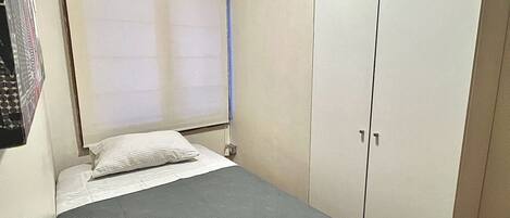 Una habitación pequeña con cama individual, colcha gris, almohada blanca, rejilla en el techo y un armario de dos puertas.