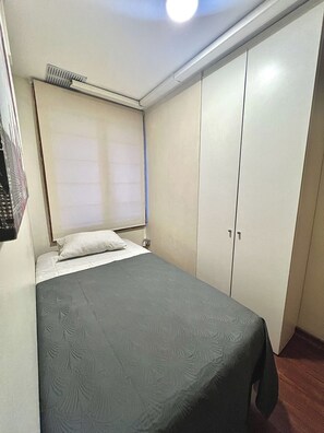 A small, single-bed room with a grey bedspread, a white pillow, a ceiling vent, and a wardrobe with two doors. - Hostel La Moneda (Santiago)