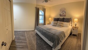 A bedroom with a large bed, two bedside tables with lamps, a window with curtains, a painting on the wall, and a patterned rug. - Down to the Wire, Cozy 2-bedroom condo with WiFi and AC in welcoming Auburn (Auburn)