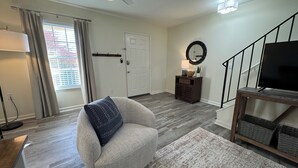 A living room with a grey armchair, a wooden floor, a staircase, a TV on a wooden stand, and a door with a coat rack. - Down to the Wire, Cozy 2-bedroom condo with WiFi and AC in welcoming Auburn (Auburn)