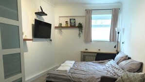 Une chambre à coucher avec un lit, une table de chevet et un petit tabouret blanc. On y trouve une fenêtre avec des rideaux, une étagère avec des plantes et un téléviseur fixé au mur.