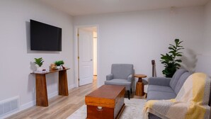 Un salon comprenant un canapé, un fauteuil, une table basse en bois et un téléviseur fixé au mur.