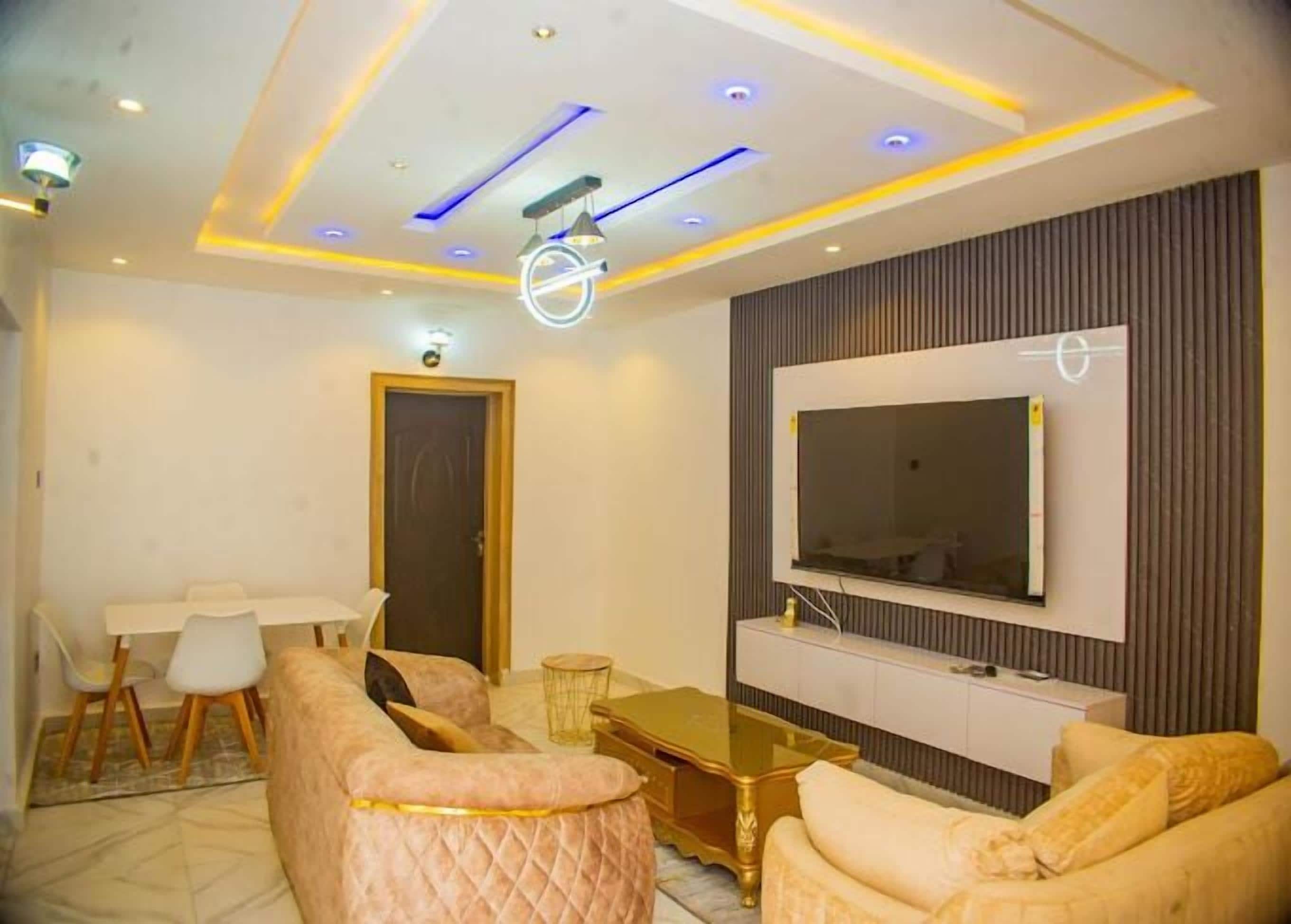 A modern living room with a large flat-screen TV mounted on the wall, a sofa, a coffee table, and a dining area with chairs.