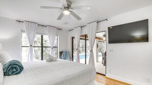 A modern bedroom with a large bed, a ceiling fan, a flat-screen TV, and a view of a swimming pool.