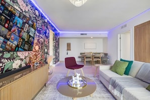Una sala de estar moderna con un mural floral, una mesa de centro redonda, un sillón rojo, un sofá con cojines verdes y una zona de comedor al fondo.