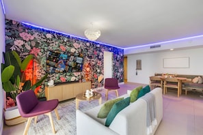 Una sala de estar moderna con papel tapiz floral, una araña, un comedor y un sofá con cojines.