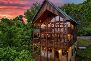 A two-story log cabin with a balcony and string lights, surrounded by lush greenery.