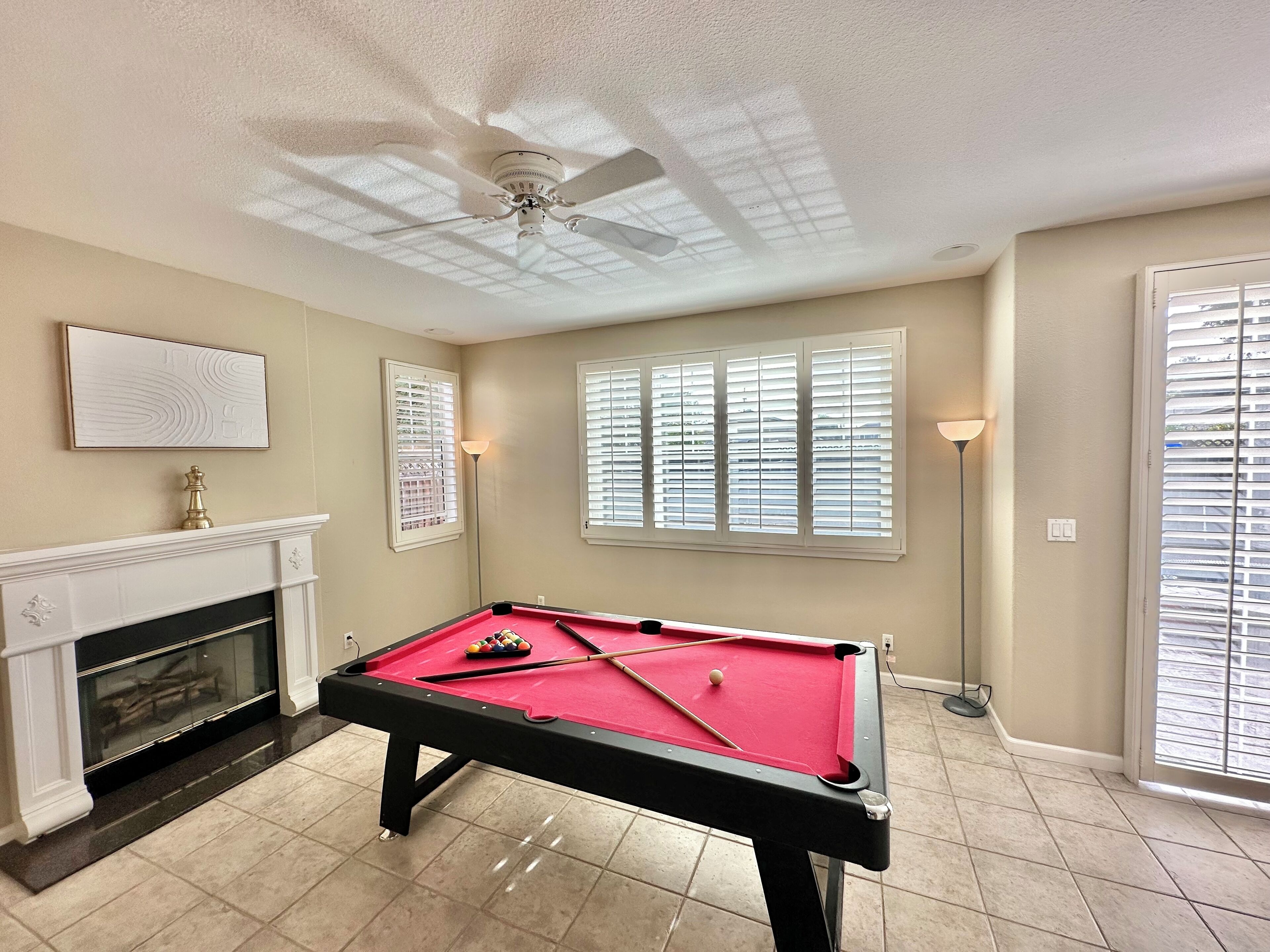 Une table de billard dans une pièce dotée d’un ventilateur de plafond, d’une cheminée et de grandes fenêtres équipées de stores.