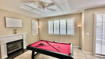 Une table de billard dans une pièce dotée d’un ventilateur de plafond, d’une cheminée et de grandes fenêtres équipées de stores.