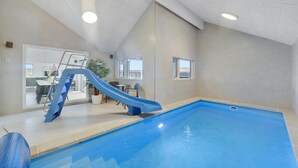 An indoor swimming pool with a blue slide, a dining area with a table and chairs, and a staircase leading to an upper level.