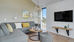A modern living room with a grey sectional sofa, a round coffee table, a grey ottoman, a TV mounted on the wall, and a window with a view of the outdoors.