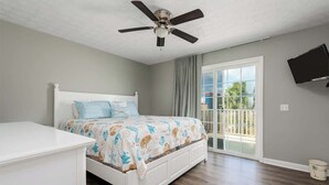 Un dormitorio con cama, ventilador de techo, televisión montada en la pared y puerta corrediza de vidrio que da a un balcón.