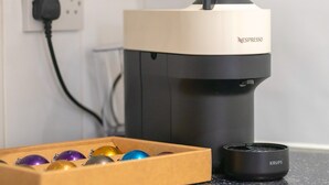 En Nespresso-kaffemaskine med en Krups-kaffekande ved siden af, en stikkontakt og en æske med farverige kaffekapsler.