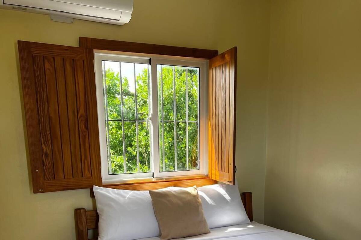A bedroom with a bed, wooden headboard, a window with a view of greenery, and a wall-mounted air conditioner.