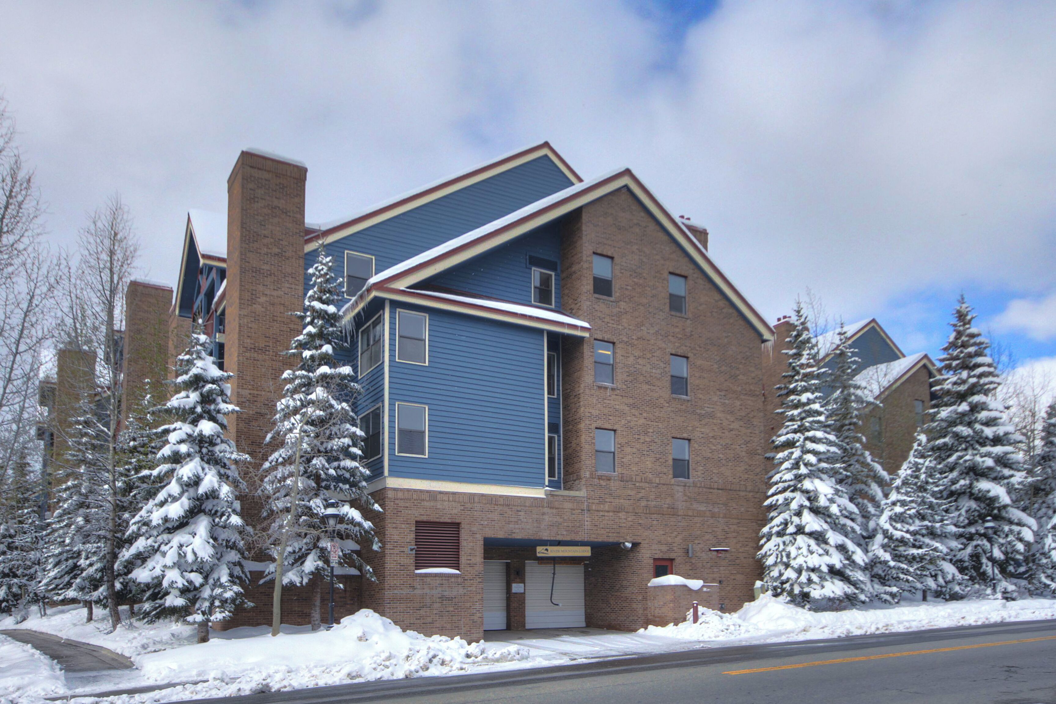 Un edificio de varios pisos con fachada azul, niveles inferiores de ladrillo y árboles cubiertos de nieve.