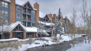 A multi-story building with a ski rental sign, a river in front, and snow-covered trees. - Downtown Breck Base Camp — Walk to Main St & Gondola — Pool + Hot Tubs (Breckenridge)