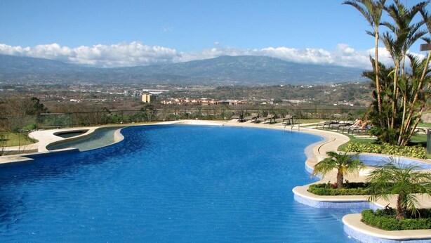 A large, clear blue swimming pool with a curved design, surrounded by palm trees and a view of distant mountains.