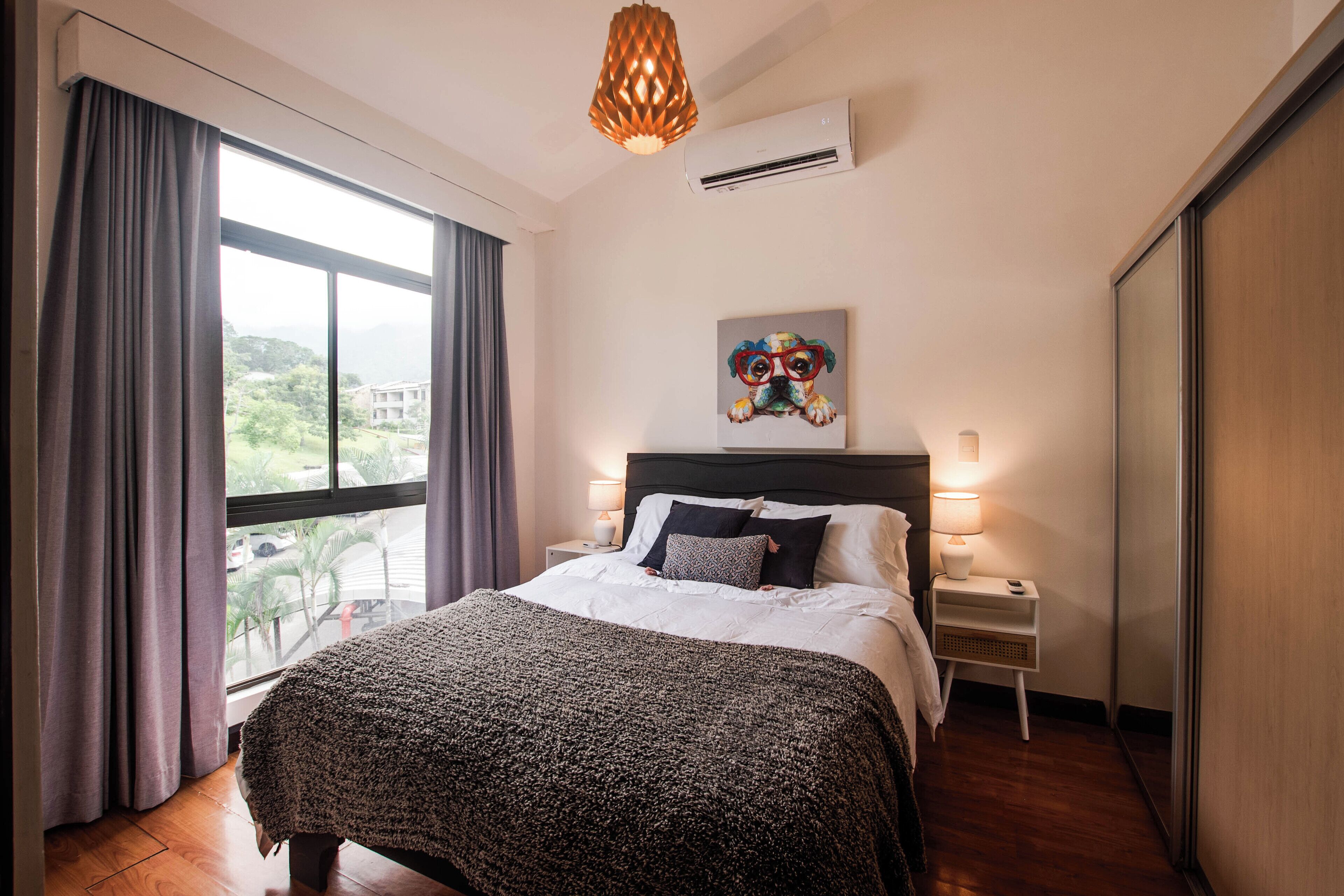 A hotel room with a large bed, a nightstand with a lamp, a painting on the wall, and a view through the window.