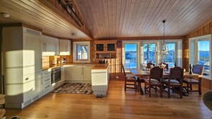 A spacious kitchen with wooden cabinets and a large island, a dining area with wooden chairs and a table, and large windows offering a view of the outdoors.