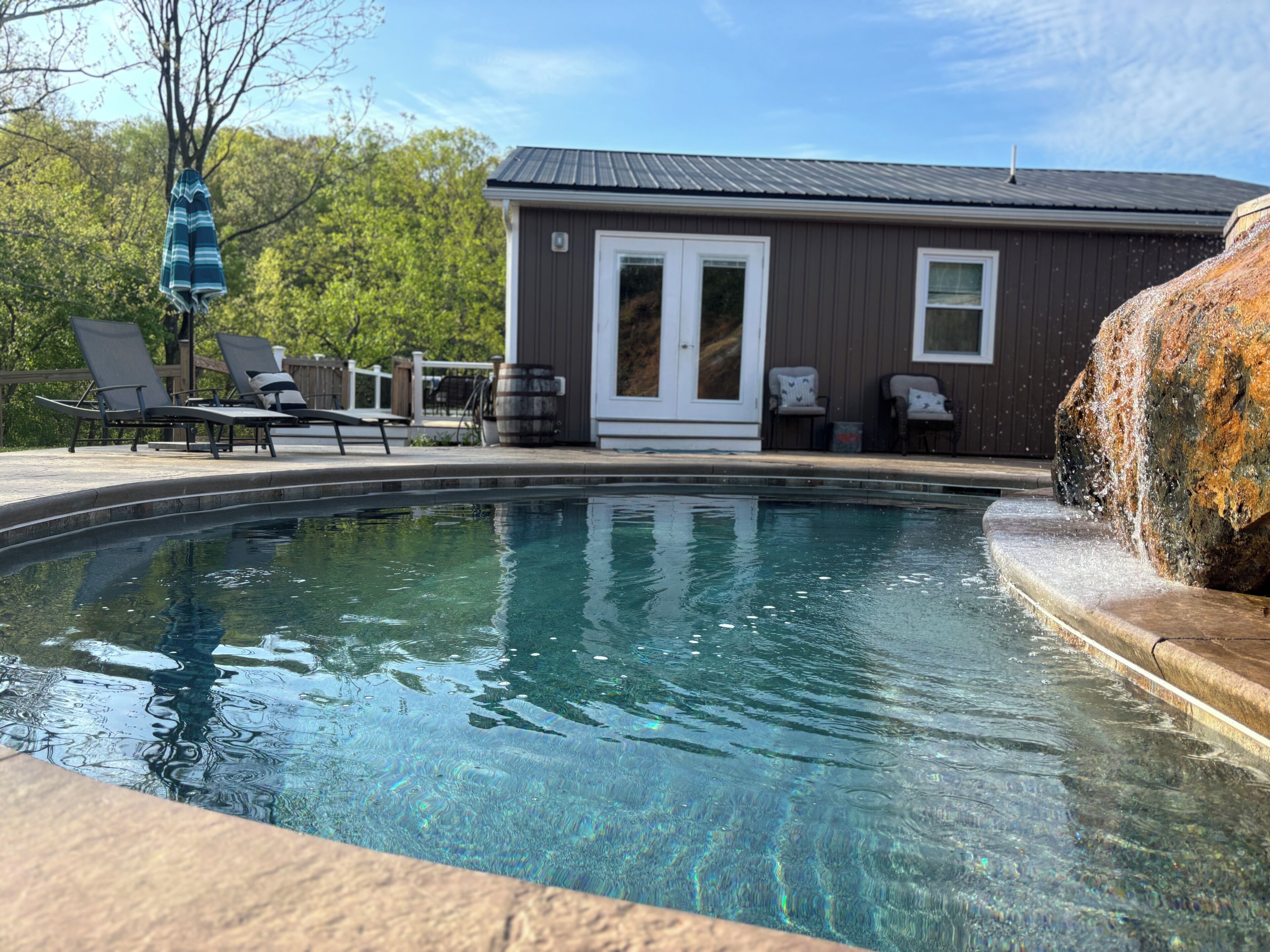Une piscine avec des chaises longues, un petit bâtiment doté d’une porte et un élément décoratif en pierre.