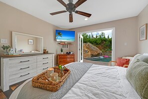 Ein Schlafzimmer mit Bett, Deckenventilator, Fernseher, Kommode, Fenster mit Blick auf Pool und Grünflächen und einem Korb mit Gegenständen auf dem Boden.