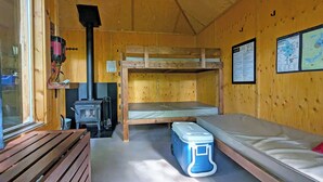 Une cabane avec des lits superposés, un poêle à bois, une carte accrochée au mur et une glacière posée sur le sol.