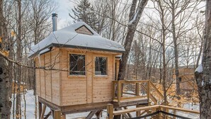 Une cabane en bois surmontée d’un toit en métal, entourée d’une terrasse en bois et d’une clôture en métal, située au cœur d’une forêt enneigée.