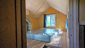 A small, single-room cabin with a sloped ceiling, a window with curtains, and a mattress on the floor.