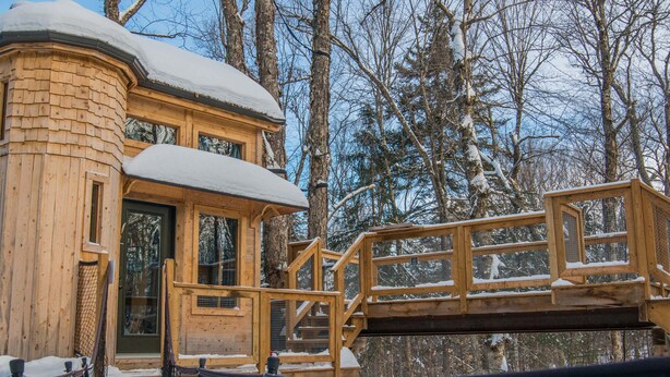 A wooden cabin with a snow-covered roof, a wooden deck, and a staircase leading to the entrance.