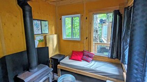 A small cabin with a single bed, a wood-burning stove, and a window with a view of trees.