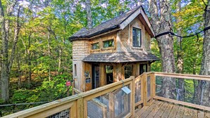 A wooden treehouse with a deck and a fenced enclosure, surrounded by a forest.