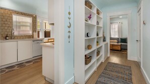 Eine moderne Küche mit weißen Schränken, einem Geschirrspüler und einem eingebauten Backofen. Ein Holzboden, ein Teppich mit Muster sowie ein Bücherregal mit Körben und Dekorationsobjekten sind ebenfalls vorhanden.