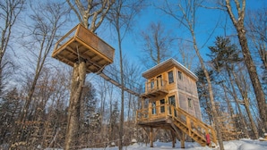 Une cabane en bois dotée d’un balcon et d’une structure en bois séparée, adossée à un arbre, le tout entouré d’une forêt enneigée.