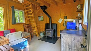 L’intérieur chaleureux d’un chalet avec un poêle à bois, un coin repas avec une table et des chaises, une fenêtre avec des rideaux et une échelle menant à un niveau supérieur.