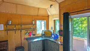 L’intérieur d’un chalet en bois comprend un coin cuisine équipé d’un poêle, d’étagères contenant des ustensiles de cuisine et d’une fenêtre donnant sur un paysage verdoyant.