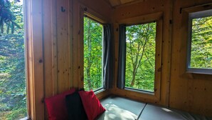 A cabin room with wooden walls, a bed with a white mattress and red and black pillows, and large windows offering a view of a forest. - Perched lakeside treehouse, suspension bridge and deck | Lodging #16 (Mont-Blanc)