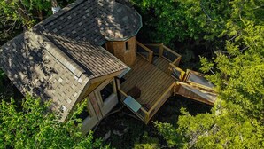 A treehouse with a wooden deck and a sloped roof, surrounded by dense foliage. - Perched lakeside treehouse, suspension bridge and deck | Lodging #16 (Mont-Blanc)