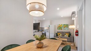 Una cuina moderna amb armaris blancs, zona de menjador amb taula de fusta i cadires verdes, i zona de sala d'estar amb sofà i un quadre.