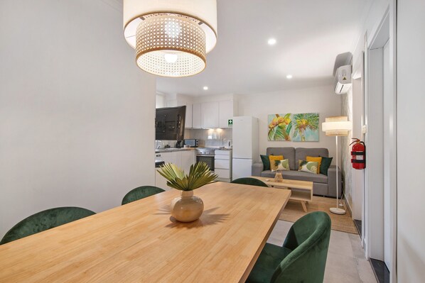 A modern kitchen with white cabinets, a dining area with a wooden table and green chairs, and a living area with a sofa and a painting.