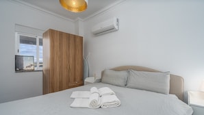 Une chambre à coucher comprenant un lit, une armoire en bois, une table de chevet, un climatiseur mural et une fenêtre donnant sur le paysage urbain.