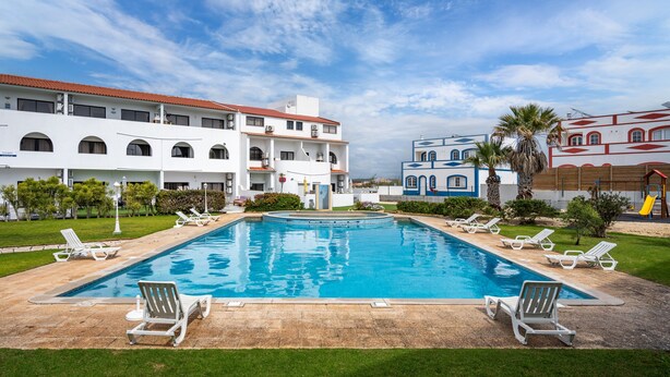 Une piscine avec des chaises longues, un bâtiment à plusieurs étages et un ciel dégagé.