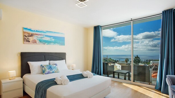 A hotel room with a large bed, a nightstand, a painting of a beach, and a balcony with a view. - Holiday Home 'Pico Do Paraíso' with Sea View, Wi-Fi and Air Conditioning (Funchal)