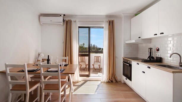 A modern kitchen with white cabinets, a dining area with wooden chairs, a large sliding glass door to a balcony, and a built-in oven.
