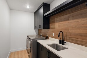A compact kitchen with a built-in washer and dryer, a sink with a black faucet, and wooden paneling on the wall. - Our Place; Our Home has an inside slide, gourmet kitchen, view for days, and just feels like home! (Hurricane)