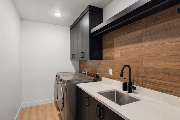 A compact kitchen with a built-in washer and dryer, a sink with a black faucet, and wooden paneling on the wall. - Our Place; Our Home has an inside slide, gourmet kitchen, view for days, and just feels like home! (Hurricane)