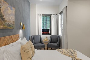 Ein modernes Hotelzimmer mit Bett, zwei Sesseln, einem Fenster mit Vorhängen, einem Wandbild und einem kleinen Tisch mit einer Vase.