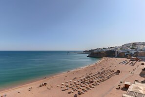 A beach with rows of sun loungers and umbrellas, a clear sky, and buildings in the background.