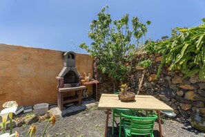 A rustic outdoor area with a stone wall, a wooden table, green chairs, a small brick oven, and a potted plant.