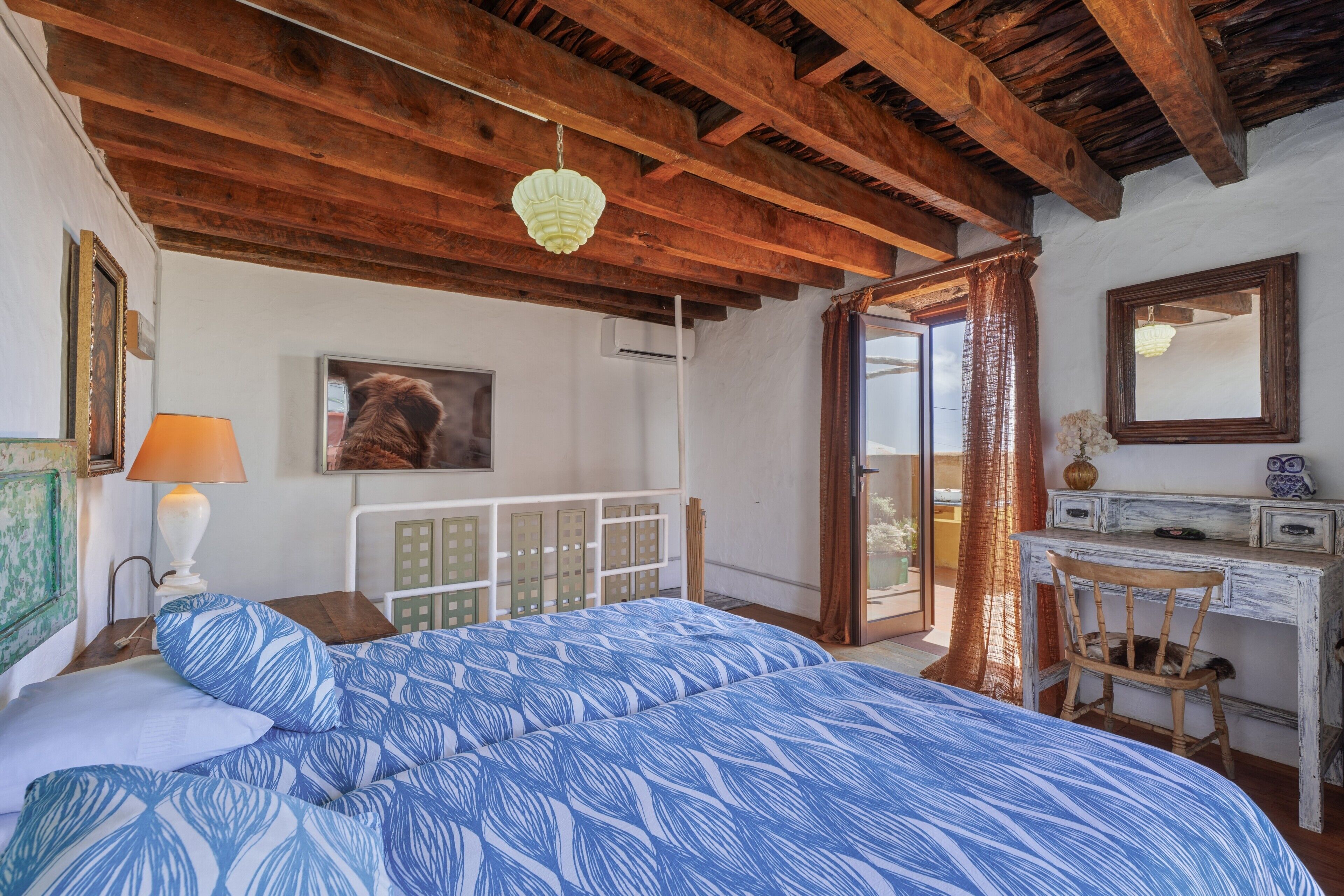 Un dormitorio con una cama, un escritorio, una silla, una lámpara colgante, un cuadro enmarcado y una ventana con cortinas.