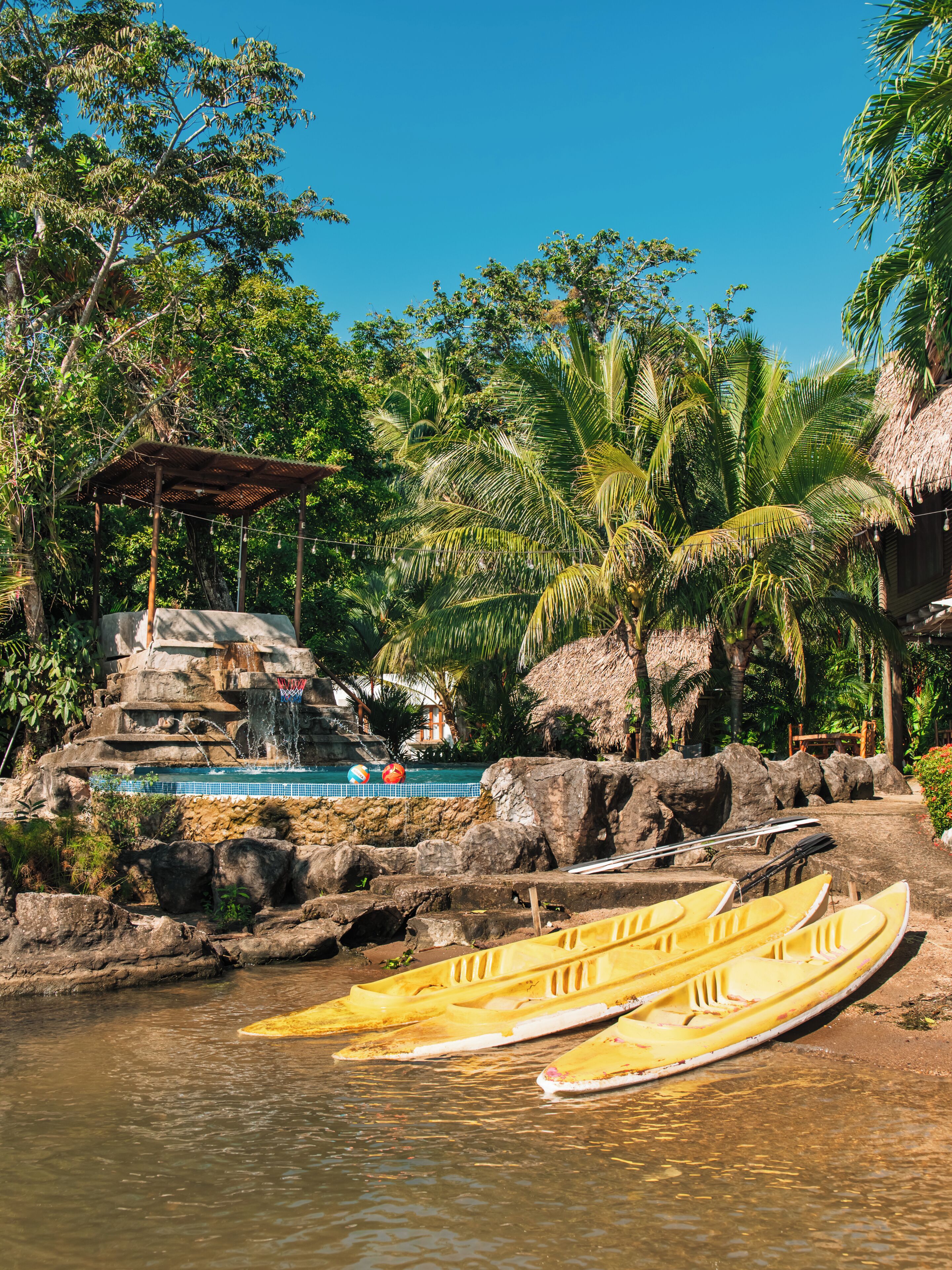 A tropical resort with a pool, thatched-roof structures, and canoes by the water.